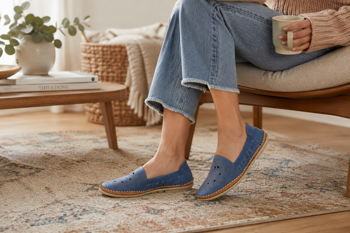 A person sitting on a chair in a cozy living room setting, wearing blue slip-on shoes with a braided jute sole. The person is also wearing light blue jeans and holding a mug.
