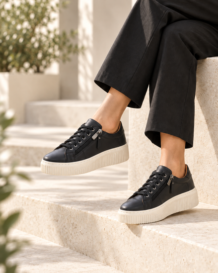 A person wearing black leather low-top sneakers with white platform soles, seated on light beige stone steps outdoors.